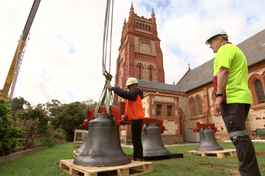 Two workers observe as a church bell is hoisted off the ground by a crane, in front of a large church tower.