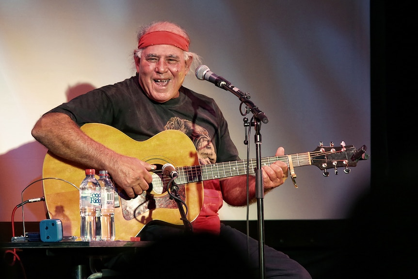 Musician Kev Carmody wearing red head band, sitting in front of microphone and laughing while on stage playing guitar.