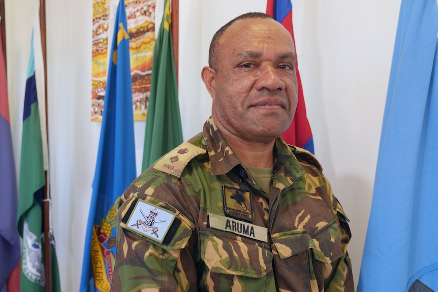 Man stands in military uniform and looks at camera with flags in background.