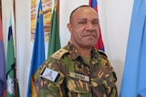 Man stands in military uniform and looks at camera with flags in background.