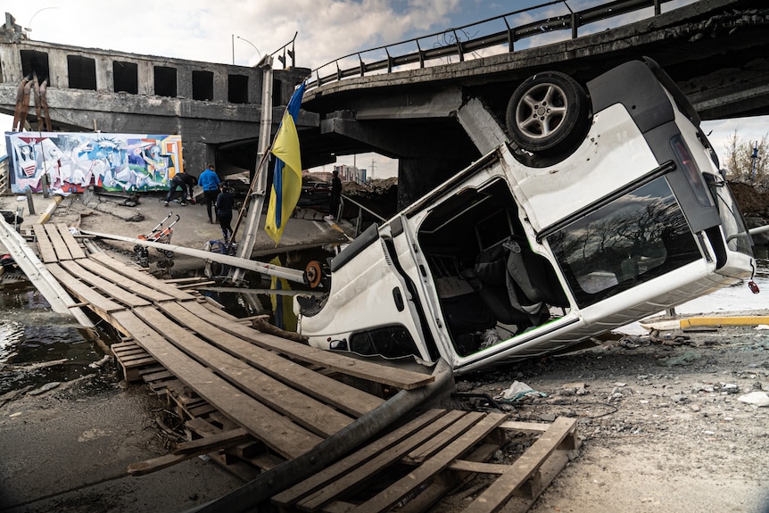 A damaged bridge, with an upturned truck next to it, and a Ukrainian flag staked in the mess 