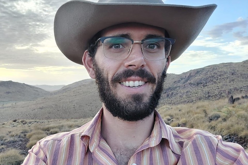 A man in a cowboy hat with a bear and glasses stands in front of hills.
