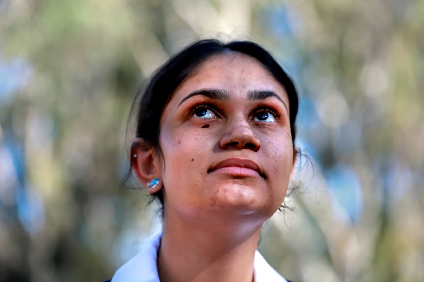 Young girl with dark skin looking upward with sun and shadow on her face and trees behind.