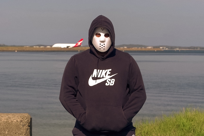 A man wearing a hockey mask stands with his hands in the pocket of his black hoodie. Behind, across a bay, a Qantas plane taxies