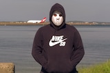 A man wearing a hockey mask stands with his hands in the pocket of his black hoodie. Behind, across a bay, a Qantas plane taxies