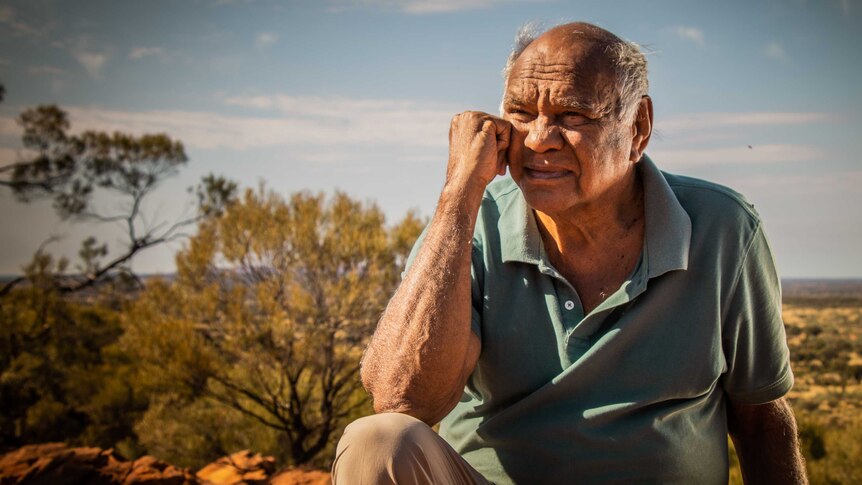 Indigenous elder sits on top of rock, in front of a  desert canyon.