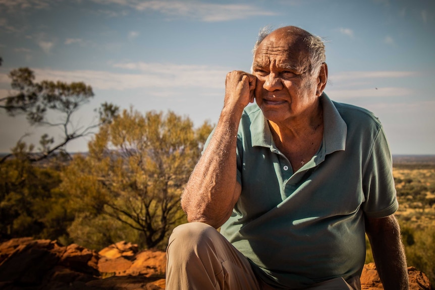Indigenous elder sits on top of rock, in front of a  desert canyon.