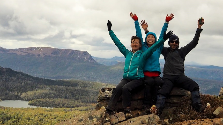 Mel and her friends wave their hands in the air, posing at a scenic lookout in the Tasmanian bush.