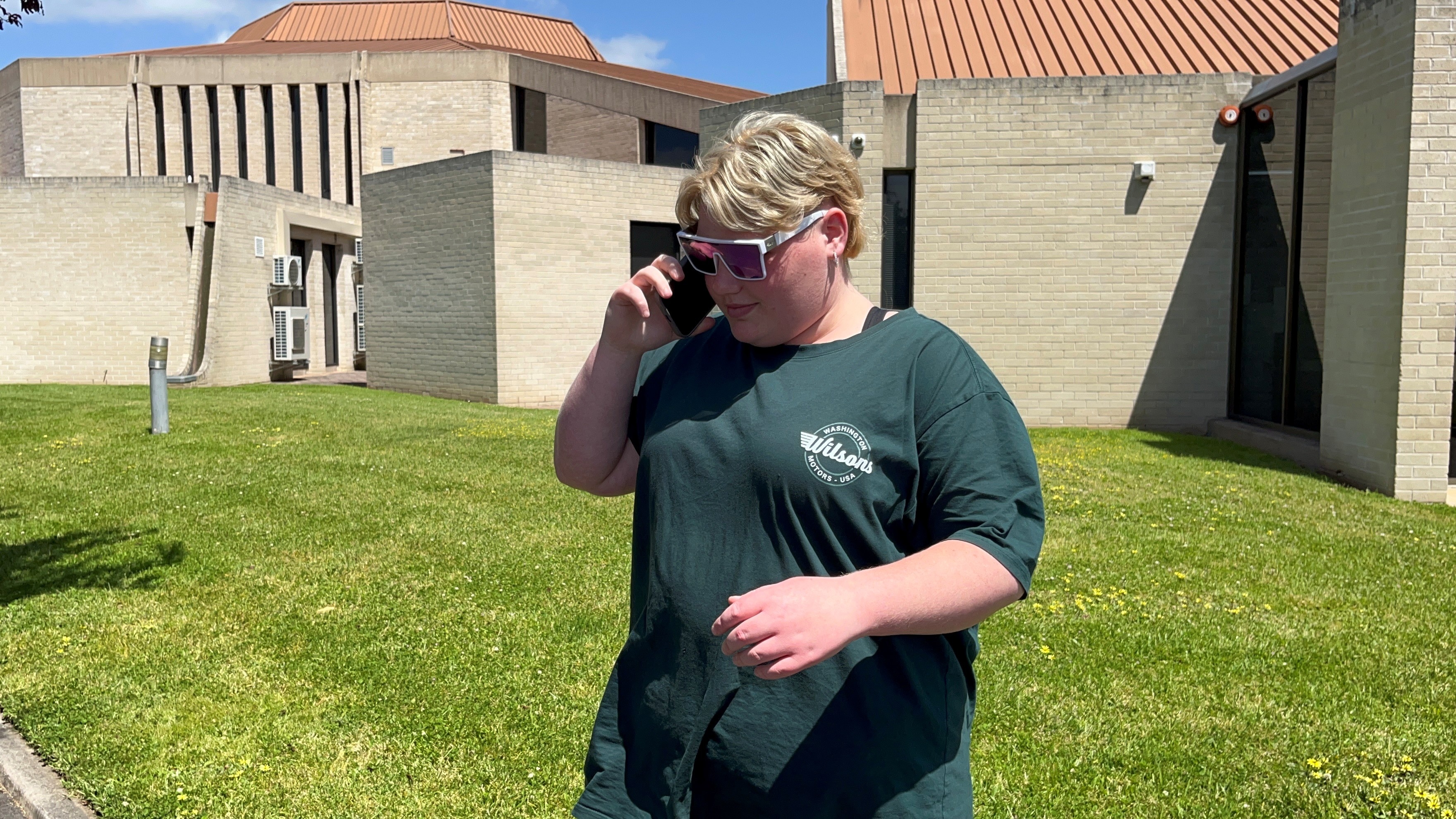 A young woman with short blonde hair wearing sunglasses and a T-shirt outside a concrete building