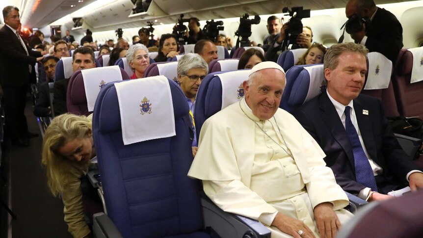 Pope Francis is flanked by Greg Burke and sits amongst journalists.