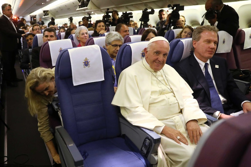 Pope Francis is flanked by Greg Burke and sits amongst journalists.