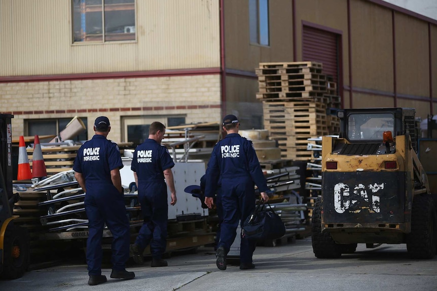 Three forensic officers stand in front of an industrial warehouse.