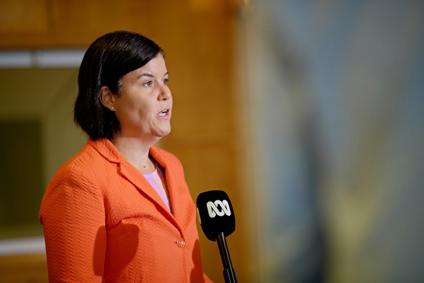 A woman in an orange blazer stands in front of a microphone.