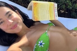 Dua smiles as she takes a selfie while lying down in a green bikini and holding a yellow book.