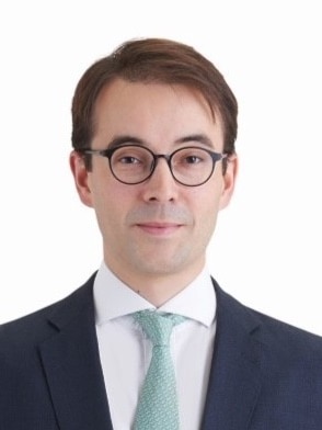 A young man in dark-rimmed glasses, suit and pale tie poses for a portrait, against a white background. 