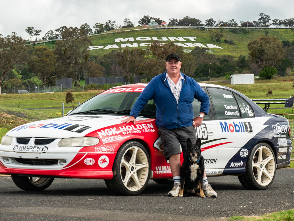 Keith Tucker standing in front of replica Holden racing car in front of Mount Panorama sign on hill.