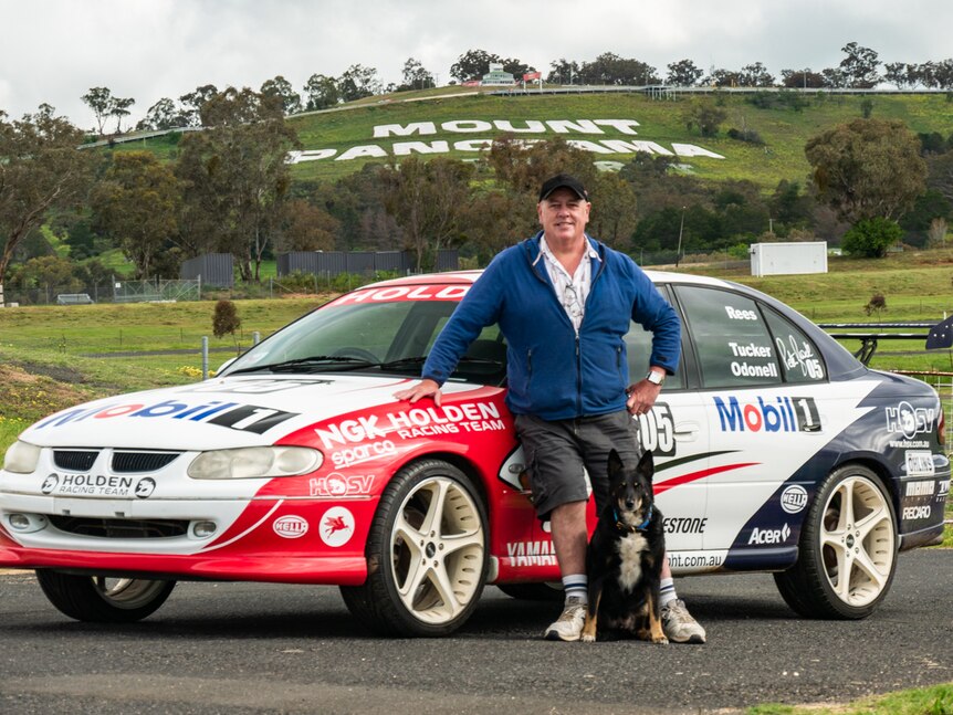 Keith Tucker standing in front of replica Holden racing car in front of Mount Panorama sign on hill.