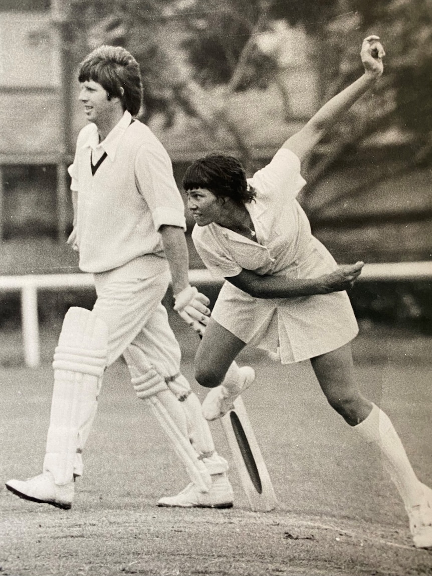A woman in cricket whites bowls as a man in cricket whites in the background prepares to run