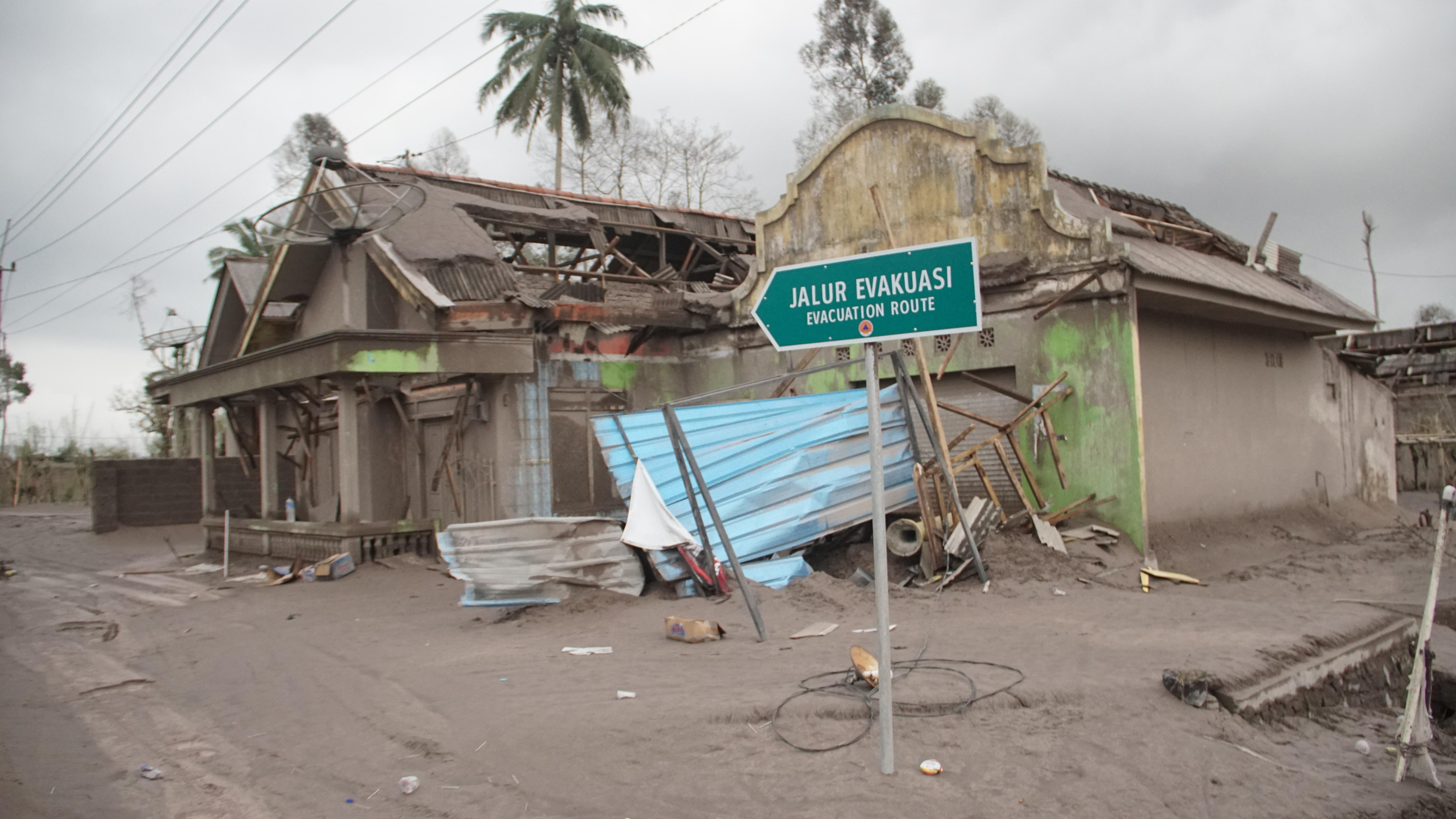 A sign post of 'Evacuation Route' in front of damaged houses covered by volcanic dusts.
