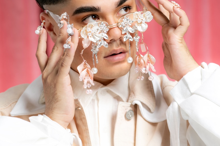 A young man looks away from the camera, wearing glasses bejewelled with crystals, pearls and shells.