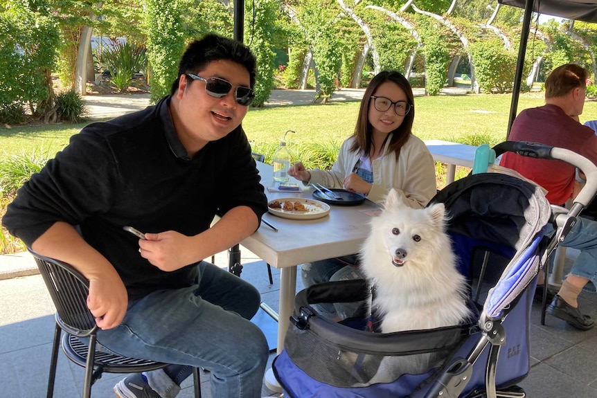 A couple sit a table at a restaurant with their dog sitting in a pram beside them.
