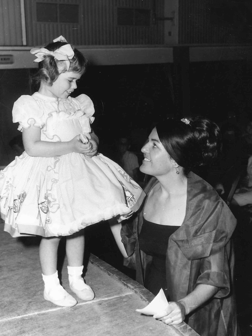 A black and white image of a small girl standing on a stage, looking down at a woman who is speaking to her. 