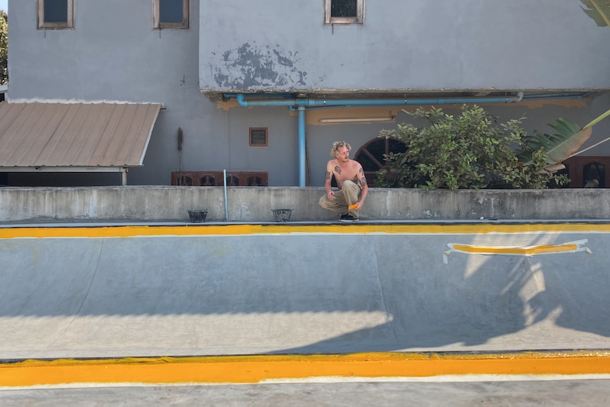 A shirtless man squatting at the edge of a concrete skate ramp in front of a house