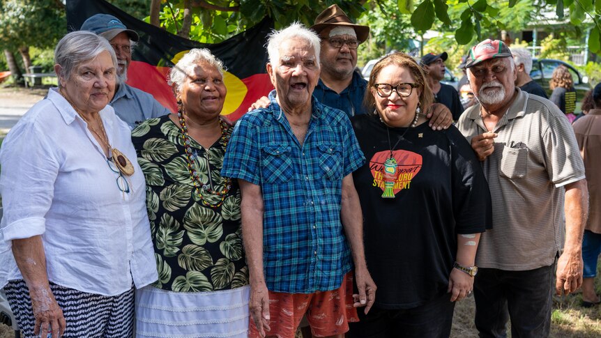 A group of First Nations leaders smile for a group photo, as someone holds the Aboriginal flag up in the background.