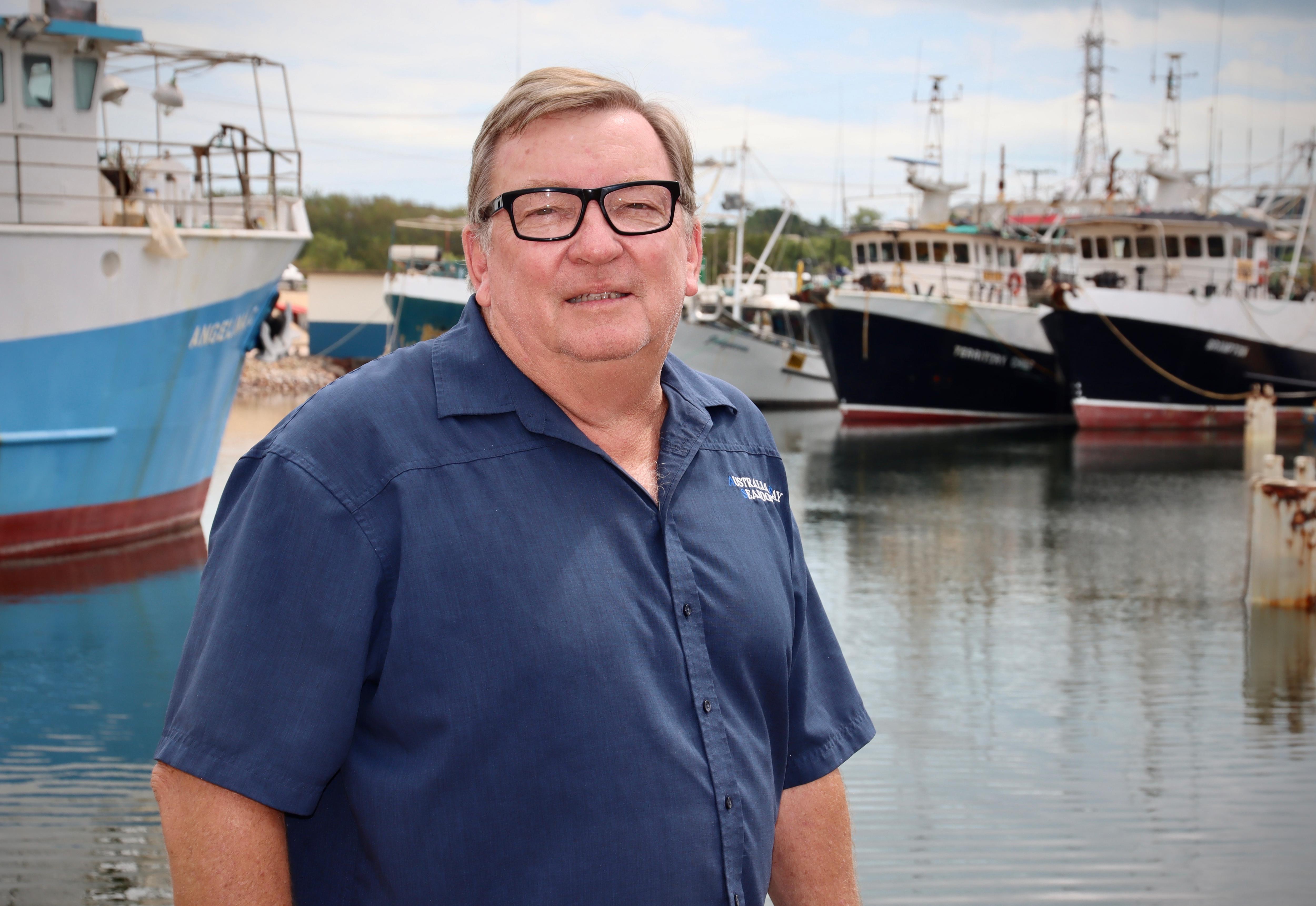 A man wearing thick, black-rimmed glasses looking at the camera with fishing boats docked behind him