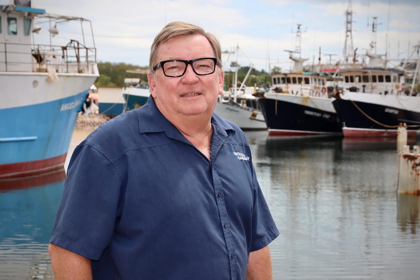 A man wearing thick, black-rimmed glasses looking at the camera with fishing boats docked behind him