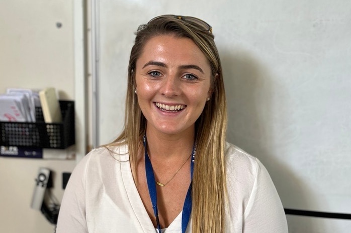 Young blonde woman smiles at camera in front of a school classroom whiteboard 