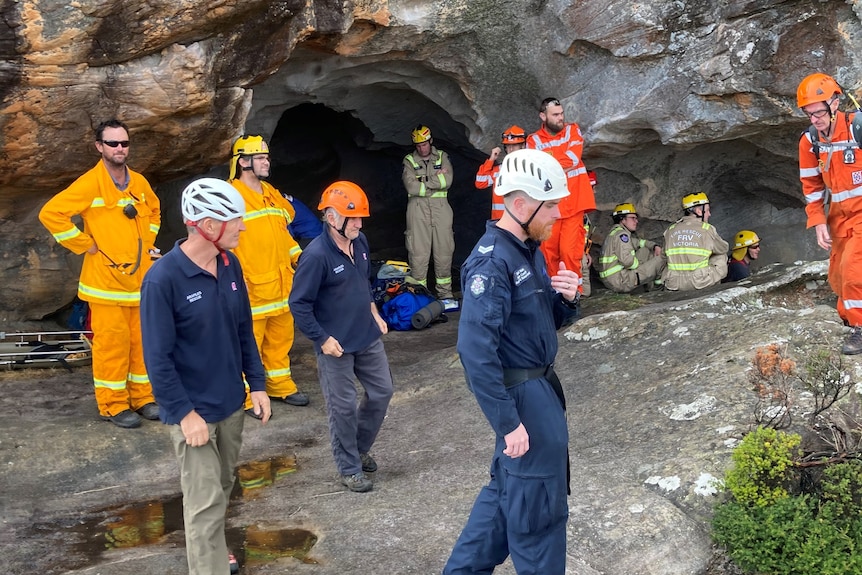A group of men in orange CFA uniform, navy police uniform and red SES uniform wearing helmets stand at the base of a cliff