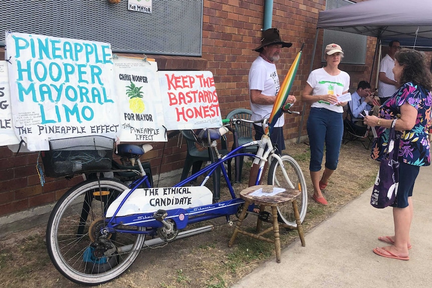 A man talks with two women. He has a bicycle and signs.