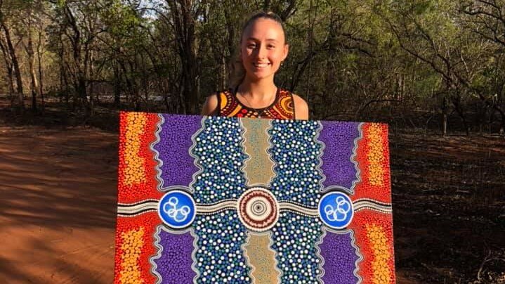 A young woman standing in bare feet in red dirt holds a colourful painting of dots and swirls