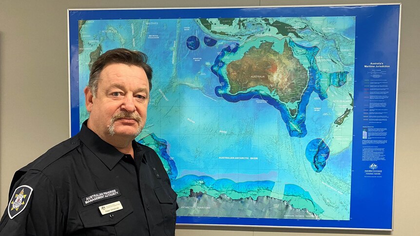 A man in a navy uniform stands in front of a large map showing Australia