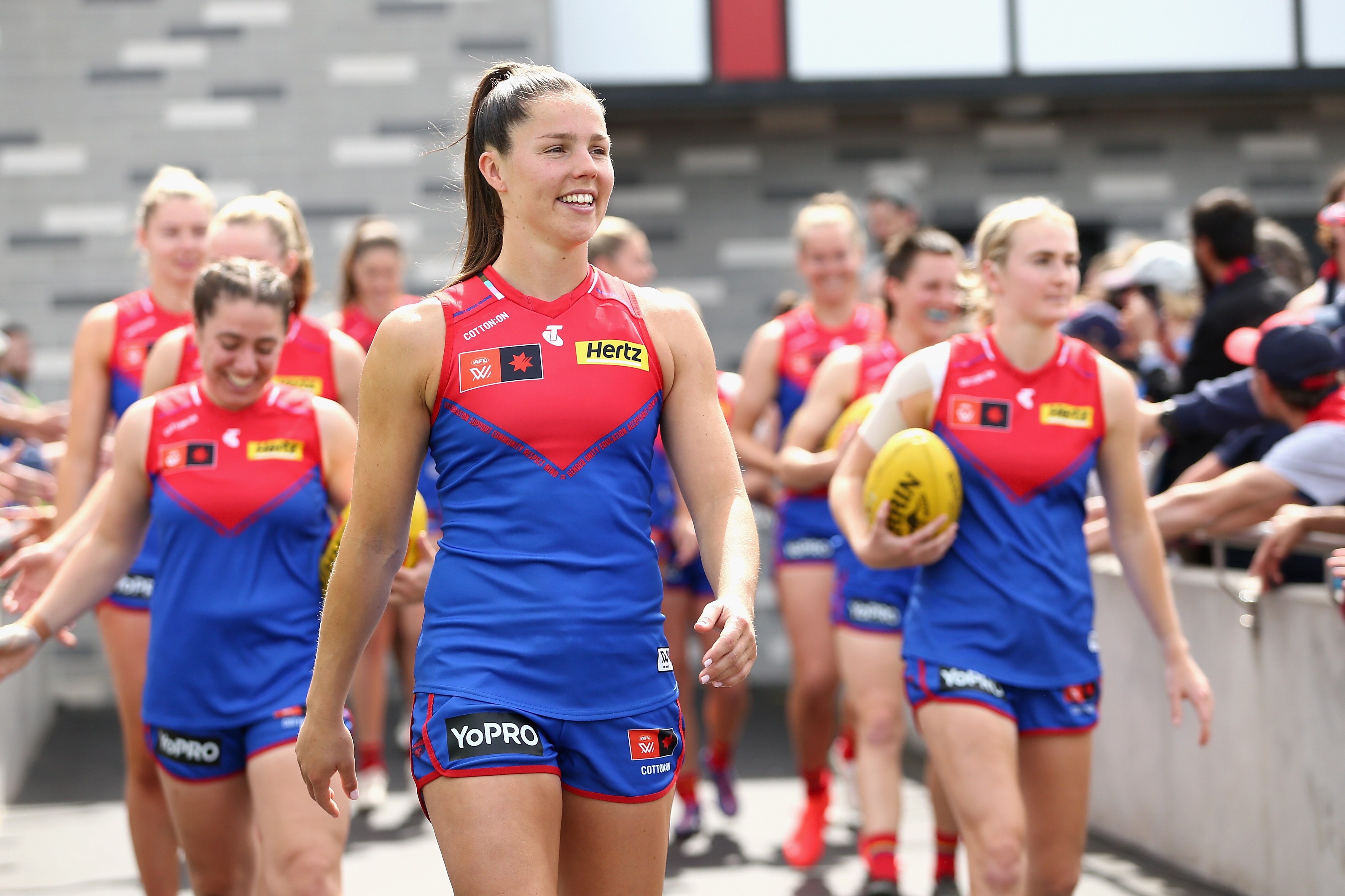 Melbourne's AFLW captain, Kate Hore, smiles as she walks on the ground leading her team behind her.