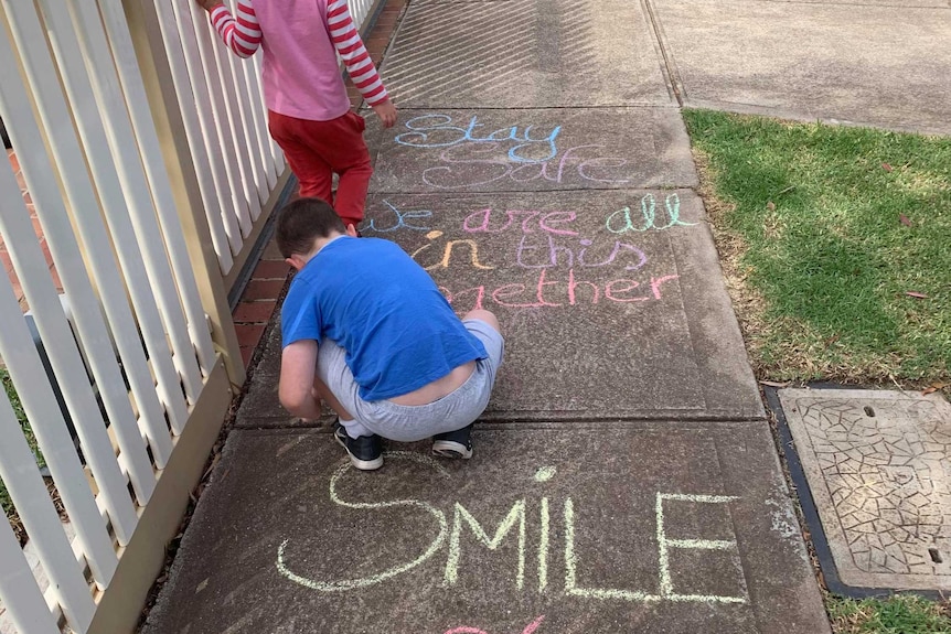 Two young children draw chalk messages like 'be happy' and 'we're all in this together'.