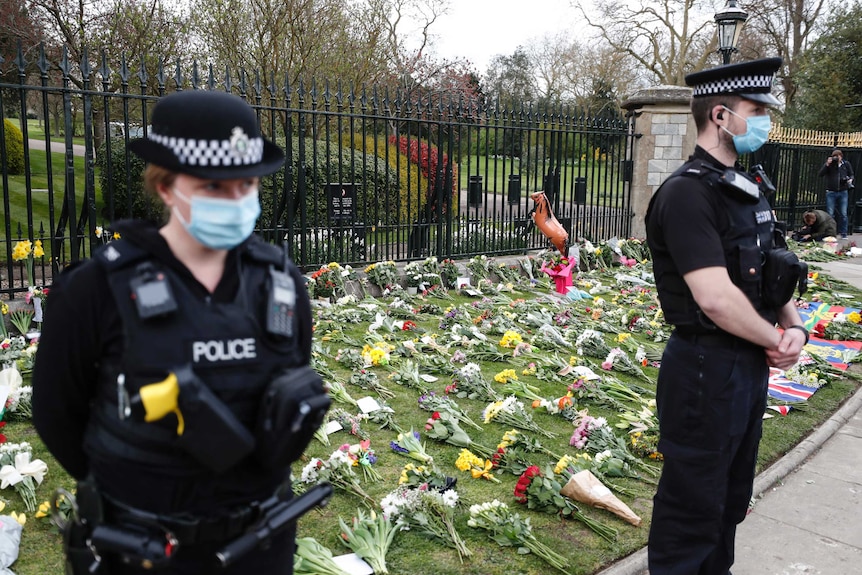 Two police officers are seen standing in front of a fence with a patch of grass behind them with approximately 50-100 bouquets.