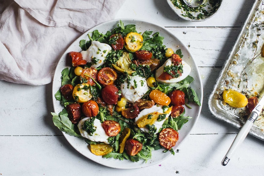 A plate of caprese salad recipe with a bed of baby rocket, topped with roasted tomatoes, mozzarella cheese, thyme and basil.