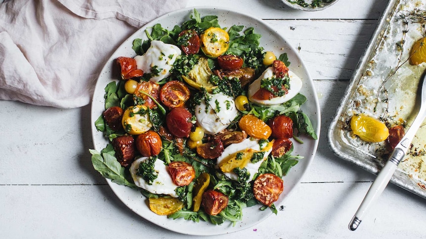 A plate of caprese salad recipe with a bed of baby rocket, topped with roasted tomatoes, mozzarella cheese, thyme and basil.