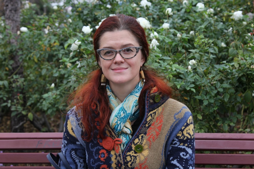 An artistic women with red hair, dangly earrings and a colourful cardigan sits on a bench in a rose garden.