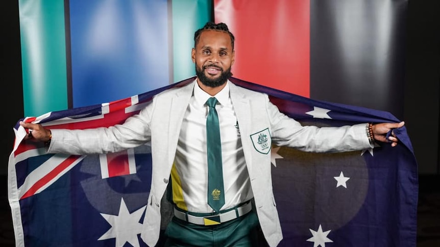 An man wearing a beige suit jacket and green pants with a green tie holds the Australia flag