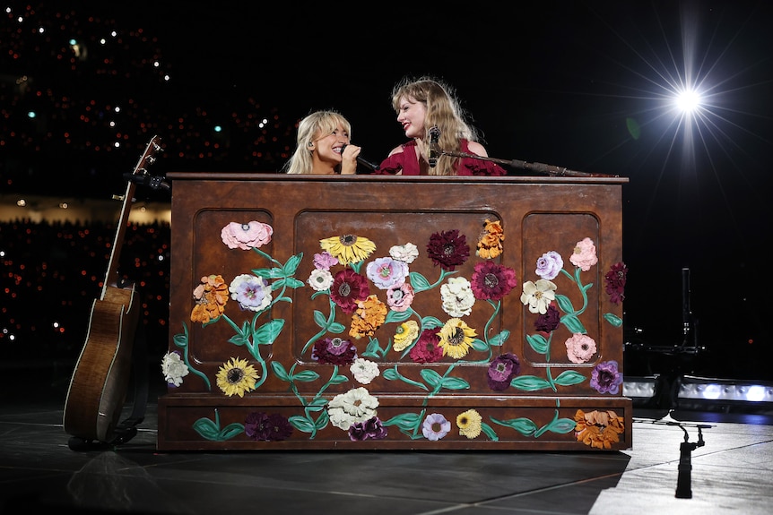 Sabrina and Taylor smile at each other on stage at a piano that’s decorated with painted flowers