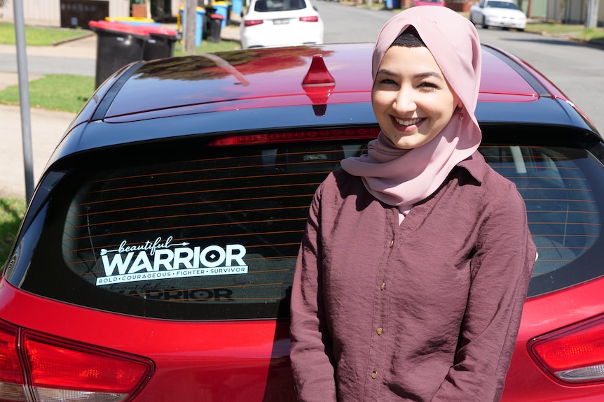 A woman standing behind her car which has a bumper sticker that says 'Beautiful warrior. Bold. Courageous. Fighter. Survivor.'