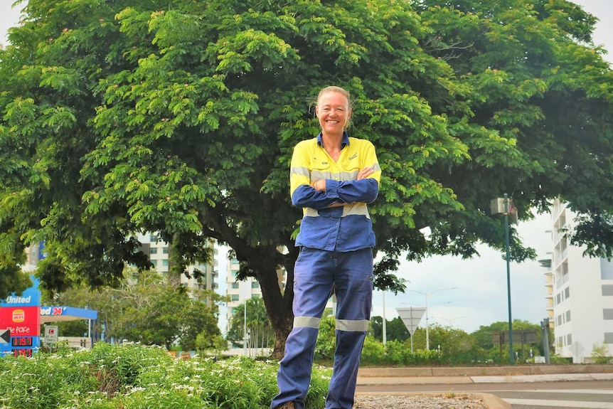 Woman in hi-viz workwear arms crossed on median strip with lush tree behind.