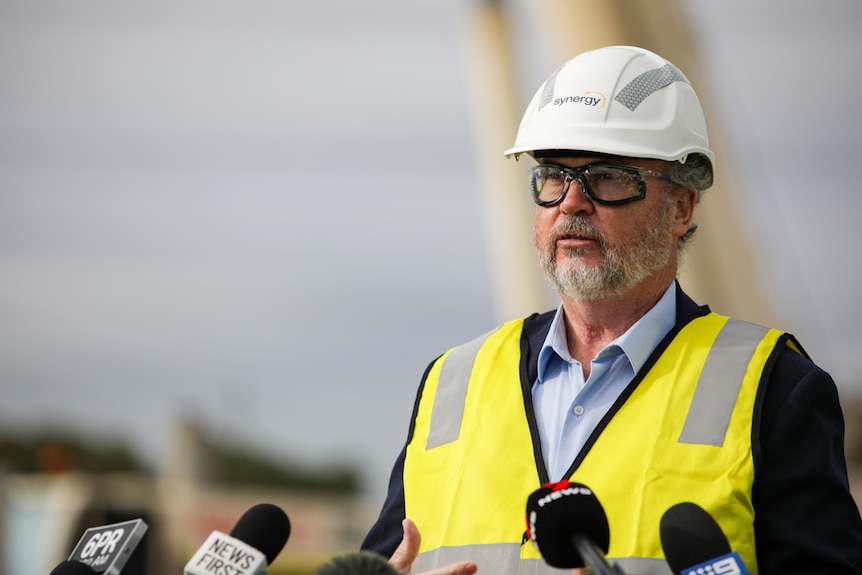 An older man, wearing a hard hat and a high-vis vest over a dark suit, speaking to the media at an industrial site.