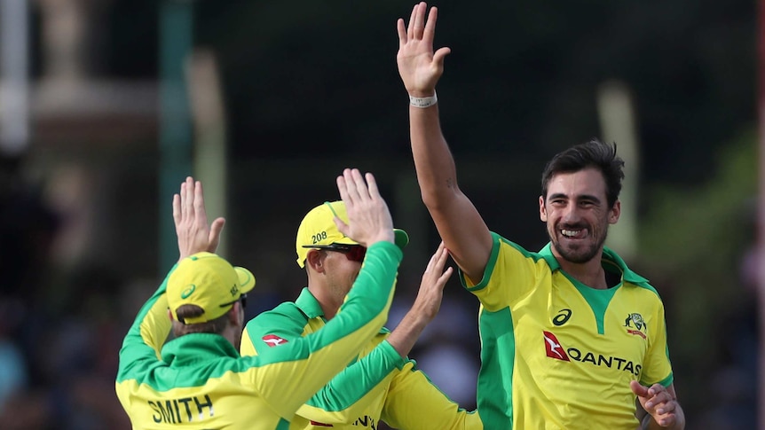 A smiling Australian fast bowler holds his hand up to high-five teammates after taking a wicket.