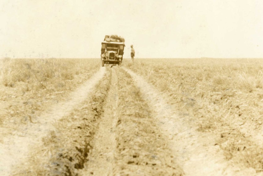 Tracks lead to a heavily laden 1930s truck on the distance in a sepia-toned photo, the surrounding land is empty.