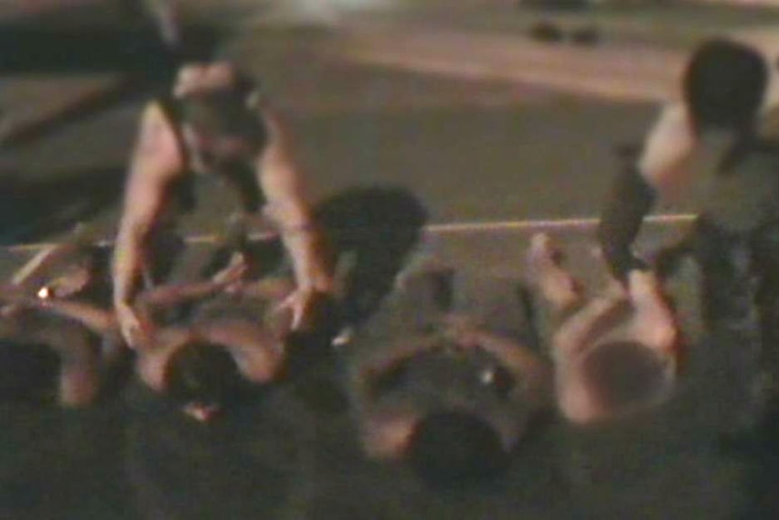 Four boys handcuffed and lying topless on the ground at night as two guards stand over them.