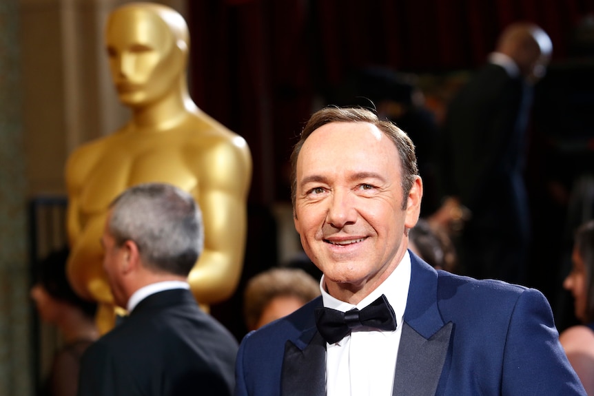 Kevin Spacey smiling in a tux, behind a giant Oscars statue 